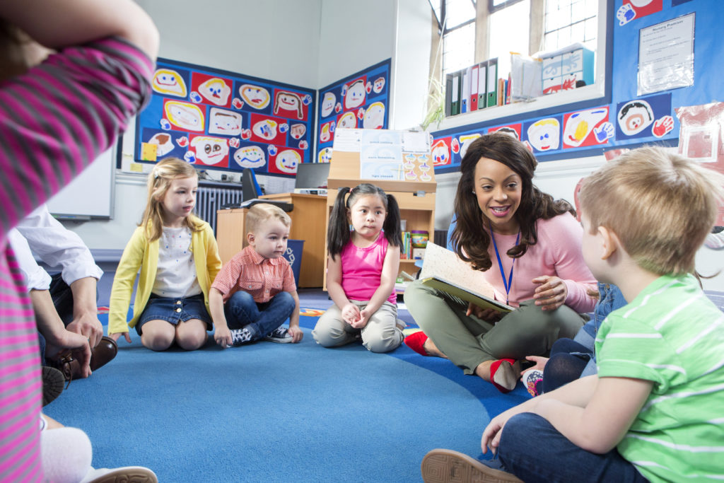 Group of nursery children sitting on the floor in their classroom. The teacher is reading from a book.